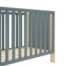 Cuna infantil de madera con acabado verde kaki y detalles en tono natural, barrotes verticales y somier de láminas de pino.
