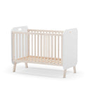 Cuna infantil blanca con barrotes verticales, laterales sólidos con asas y patas inclinadas de madera clara.