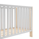Cuna infantil blanca con barrotes verticales y estructura de madera clara, de diseño sencillo y somier de láminas.