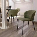 Silla de comedor tapizada en verde oliva texturizado, con respaldo curvo envolvente y patas finas de hierro negro.
