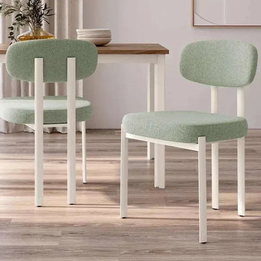 Silla de comedor tapizada en tela verde claro con patas blancas de metal, diseño moderno y respaldo ovalado acolchado.