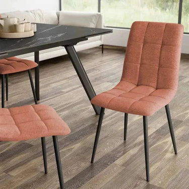 Silla de comedor con tapizado texturizado color salmón, respaldo y asiento capitoné y patas metálicas negras.